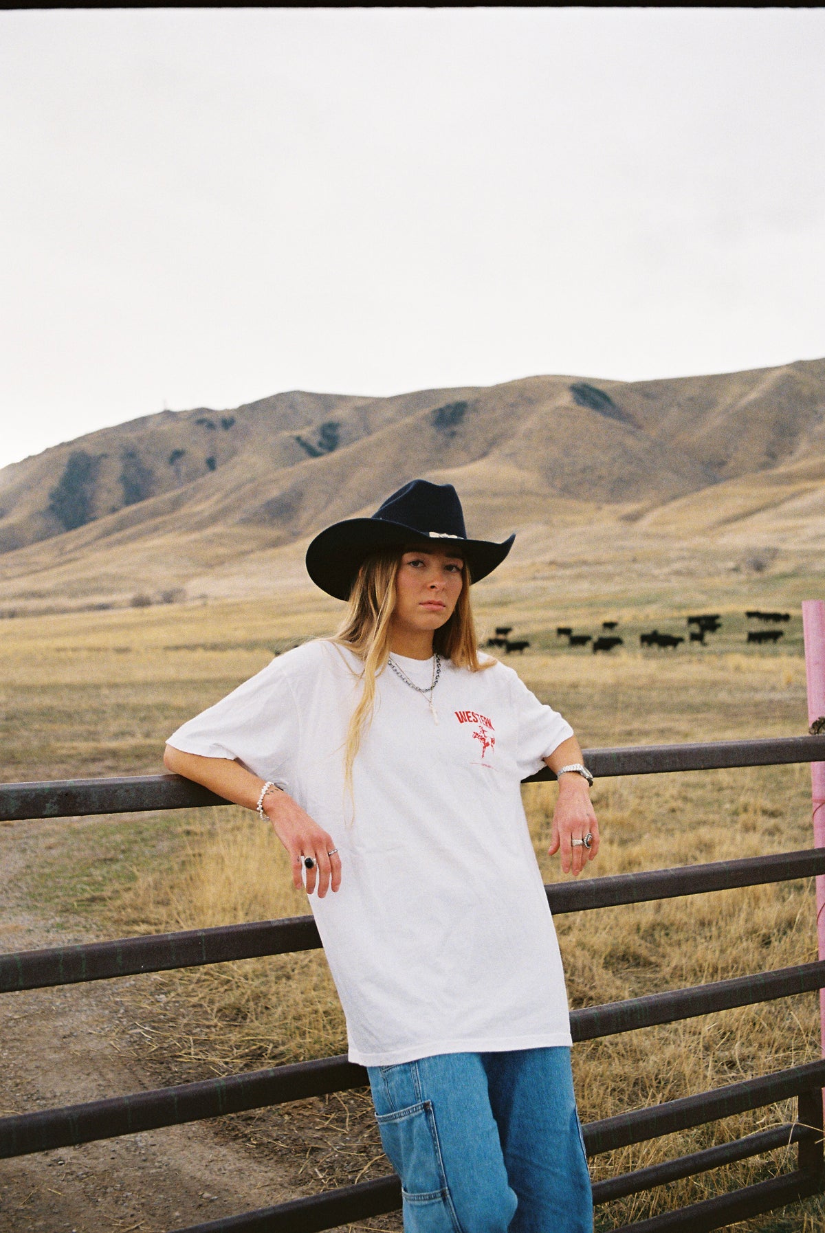 A woman wearing a white graphic t-shirt, blue jeans, and a black cowboy hat leans against a metal fence in a dry grass field with mountains in the background.